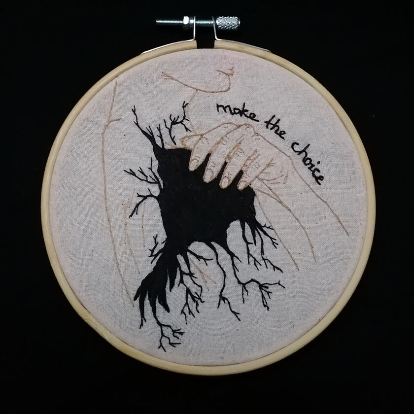 Photo of embroidery hoop. The embroidery shows a woman's shoulder with a black mass with tendrils growing out